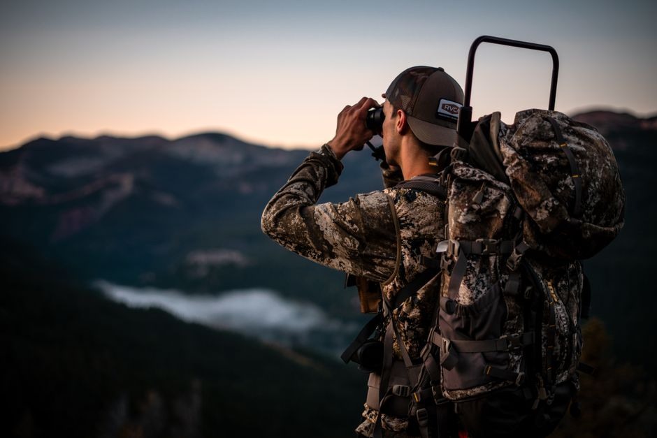 Photographer using binoculars from a rocky viewpoint in warm outdoor light.